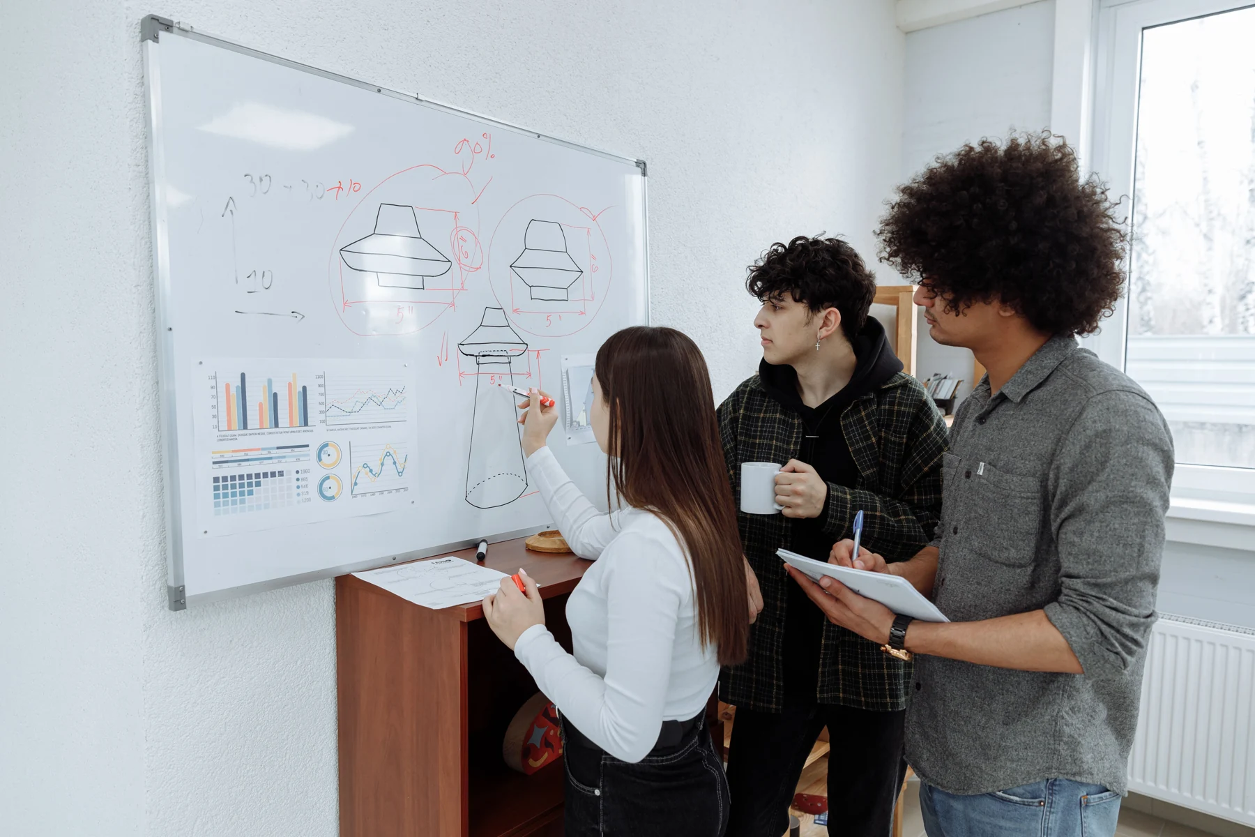 Three people discuss product sketches at a whiteboard; one draws while another takes notes.